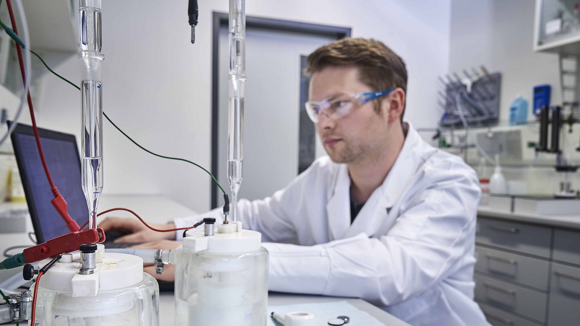 A man with safety goggles and a lab coat sits in a laboratory